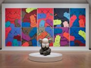Installation view, KAWS: WHAT PARTY, Brooklyn Museum, February 26, 2021 - September 5, 2021. Features sculpture in front of a series of several flat images of 