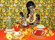 Miss meatface in neutral oranges and a leopard-print latex mask, sits in front of table loaded with pineapples, jello molds, and sausage plates. In front of busy floral background. 