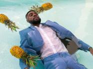 A man with dark skin, wearing a suit floats in a pool amongst three pineapples. 
