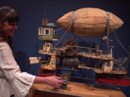 Woman turns handle of steampunk blimp ship sculpture