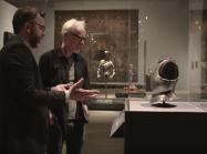 Adam Savage and Met Curator look at child's breastplate in glass case