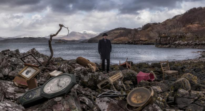John Akomfrah, Video still from Vertigo Sea, 2015. A man in old timey garb stands on the shore of a pond littered with clocks.