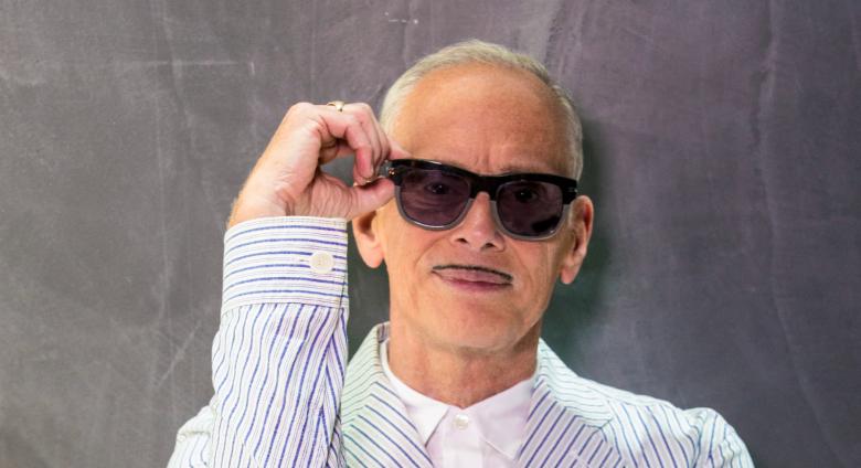 photograph of john waters, an older white man with a small moustache, wearing sunglasses and a seersucker suit