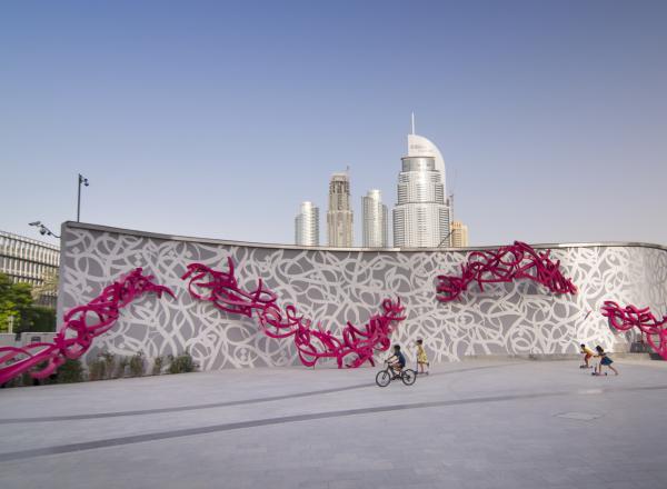 eL Seed, Declaration. Courtesy of the artist. a large installation piece in context—with city visible in the distance and children riding on bikes in front. 