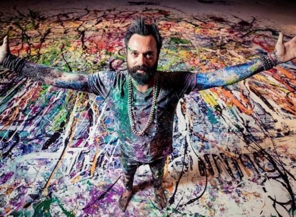 Artist Sacha Jafri covered in paint with arms spread, standing on a large paint-covered canvas