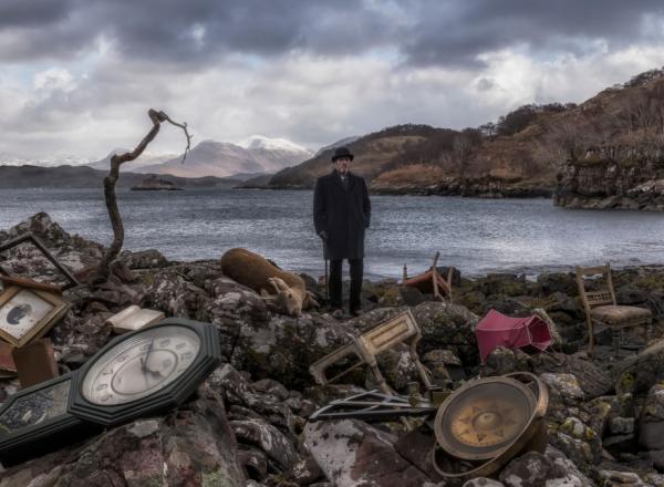 John Akomfrah, Video still from Vertigo Sea, 2015. A man in old timey garb stands on the shore of a pond littered with clocks.