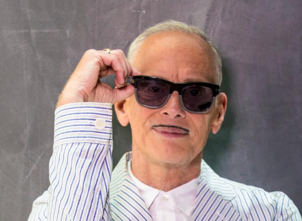 photograph of john waters, an older white man with a small moustache, wearing sunglasses and a seersucker suit