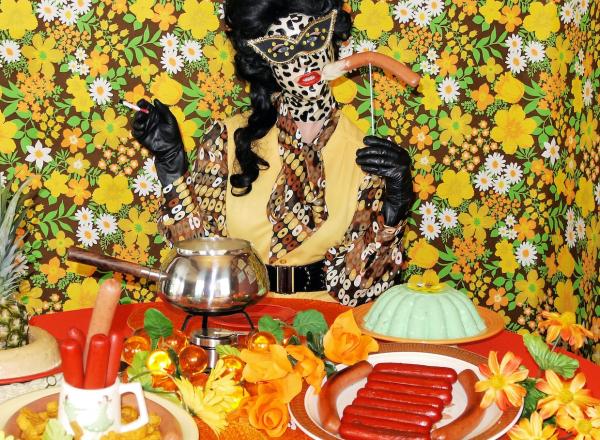 Miss meatface in neutral oranges and a leopard-print latex mask, sits in front of table loaded with pineapples, jello molds, and sausage plates. In front of busy floral background. 