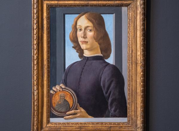 Botticelli's painting of a young man looking directly at the viewer hangs on a grey wall, in a gold frame. 