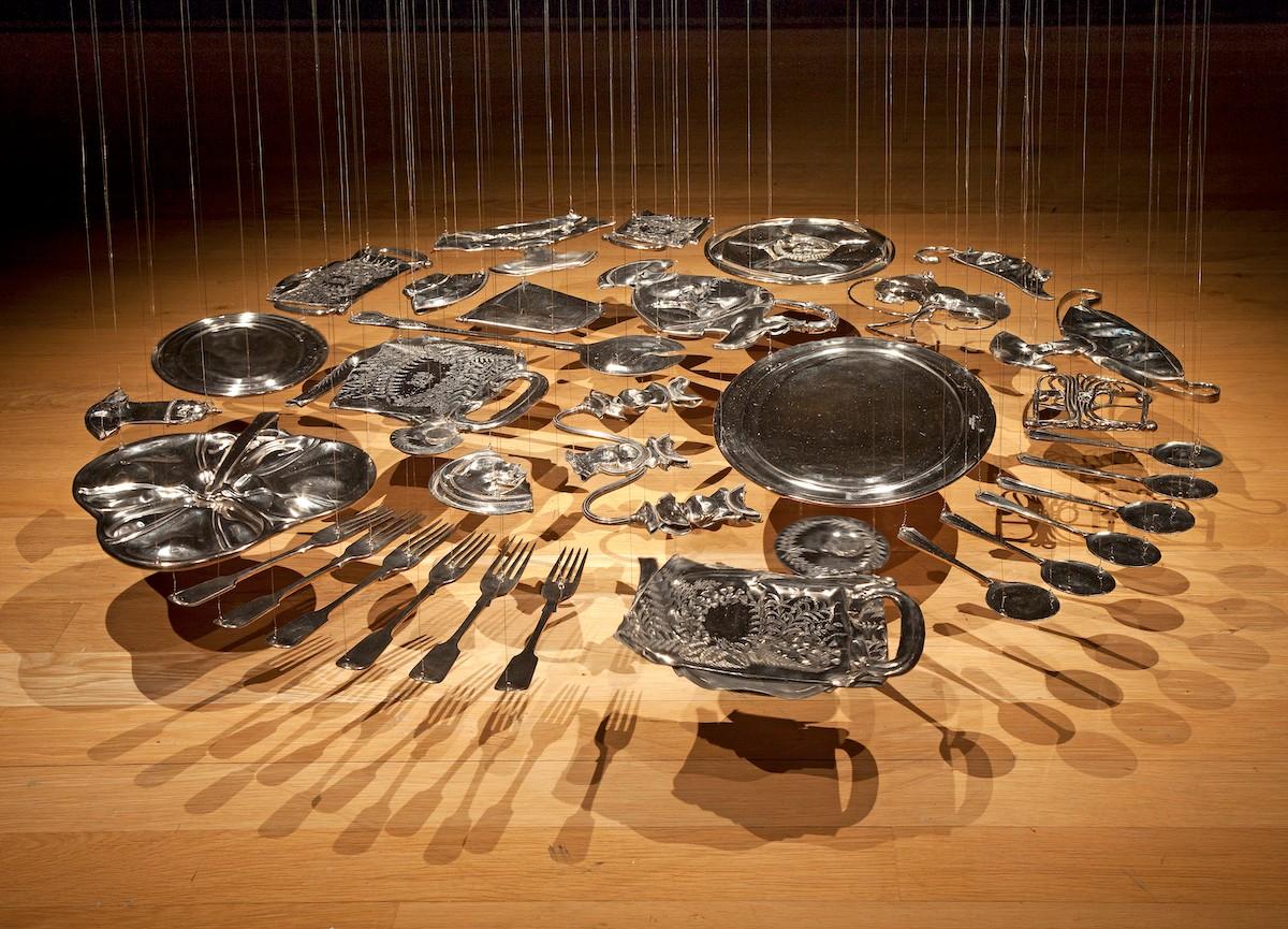 Cornelia Parker, Thirty Pieces of Silver (exhaled) Sugar Bowl, 2003; Thirty silver-plated items, including spoons, forks, trays, and various serving ware, are flattened and hang suspended from the ceiling with thin, nearly invisible wire. The pieces are arranged in a circle several inches above the floor.