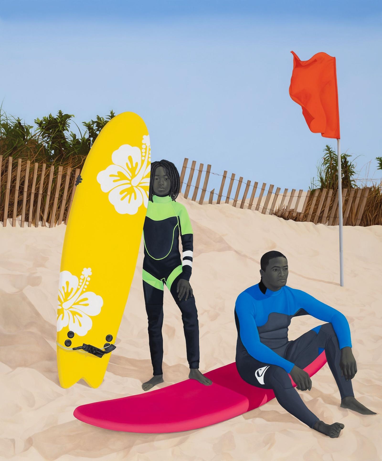 Two surfers pose, one stands with her board, looking at viewer while the other sits on his, looking out at the sea.