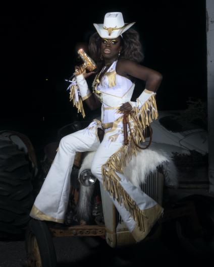 Bob dressed in fringe-covered chaps, a vest, arm-guards, and a cowboy hat. Bob holds a rhinestoned hairdryer like a gun. 