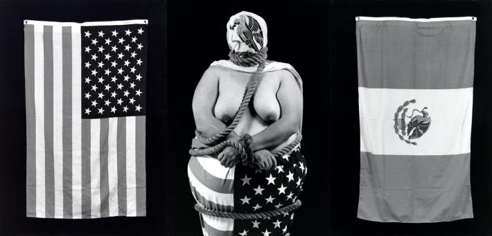 woman, bare-chested with face covered by mexican flag and legs covered by american flag stands in the middle of two more of these flags.