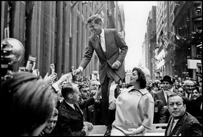 Cornell Capa photograph of JFK standing on a platform shaking hands with men in the crowd below him with Jacqueline Kennedy looking up at him