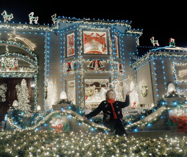 a woman in a christmas sweater sits in front of her large home decorated in christmas lights