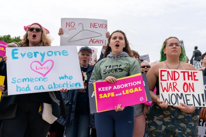 Group of protesters with signs about women's rights and abortion access. A woman in the back has a sign with a coat hanger drawn on it.