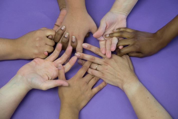 SUSAN MEISELAS photograph of hand of all skin tones intertwined on a lavender tabletop