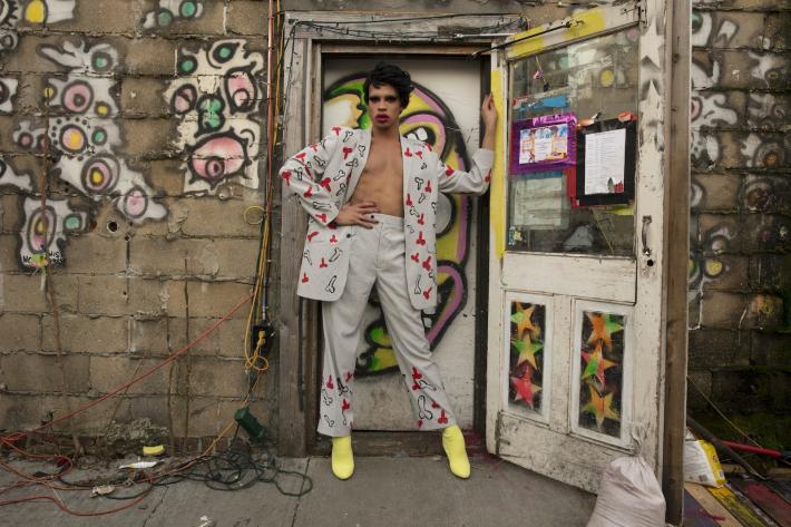 Yvie Oddly in a striped suit, covered in painted cartoon penises. She's standing in the doorway of what appears to be the back of a building with murals and fliers. 