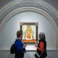 Two museum visitors are viewing a painting. The painting features a woman with a medium skin-tone wearing a dress and scarf and holding a piece of paper. It is framed by an arc with golden and floral ornamentation.